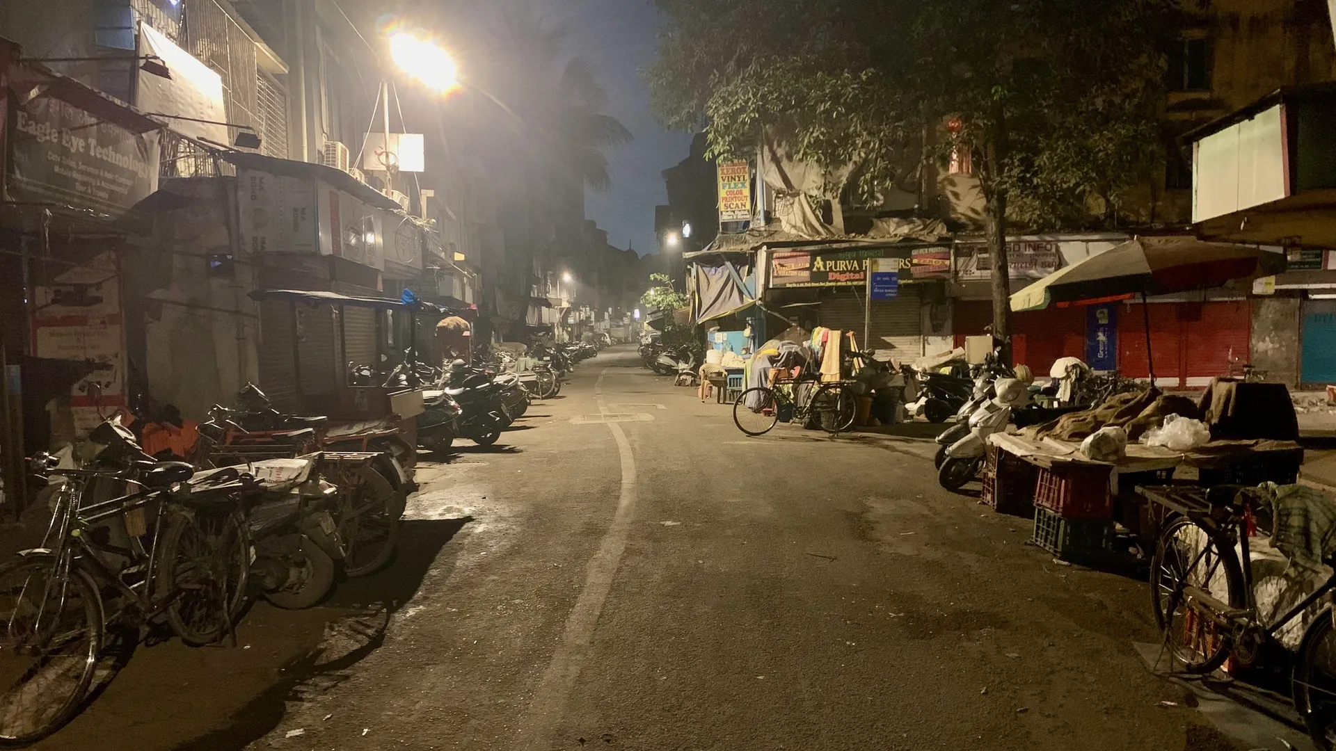 An old Mumbai bazaar lane at night — shuttered shops and parked bikes under sodium streetlight haze