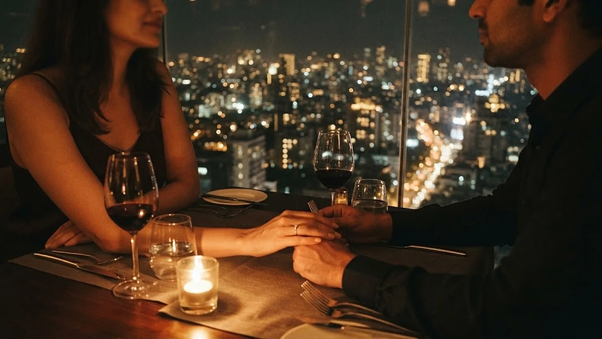 A couple holding hands over a candlelit dinner table, high above Mumbai, the city lit up beyond the window