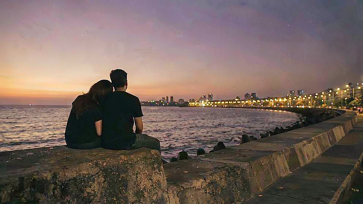 A couple seen from behind on the Marine Drive sea wall at dusk, the Queen's Necklace curving away beyond them